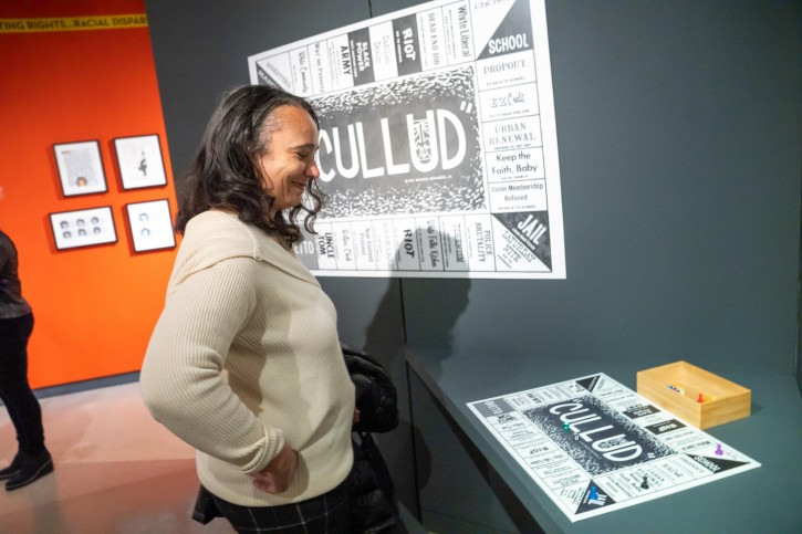 A person in a white sweater smiles while looking down at a piece of art about resembling a game board.