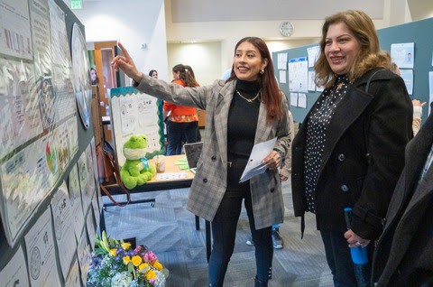 MA student gestures at academic poster while smiling woman looks on