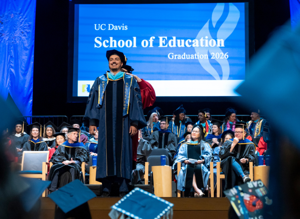 A doctoral student crosses the graduation stage. Faculty are seated in chairs behind him.