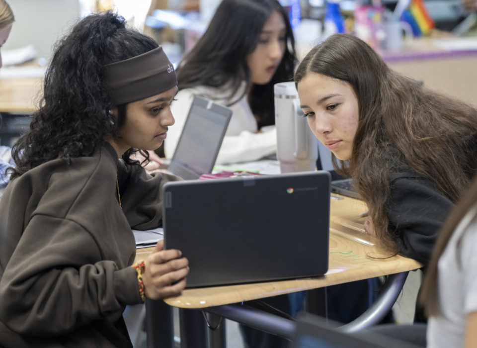 Two students sit at a desk cluster and look at a laptop screen with intensity.