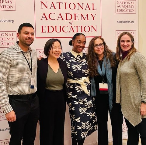 Five people pose in front of a poster for the National Academy of Education Annual Meeting & Fellows Retreat.