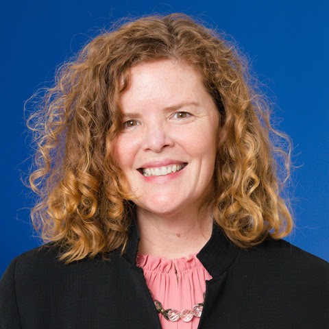 A headshot of Susanna Cooper wearing a pink shirt and a black blazer against a blue background.