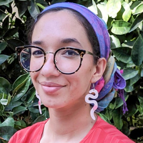 A headshot of Morgan Myhre wearing a pink shirt, a colorful purple head scarf, white snake earrings, and tortoiseshell glasses against a background of greenery.