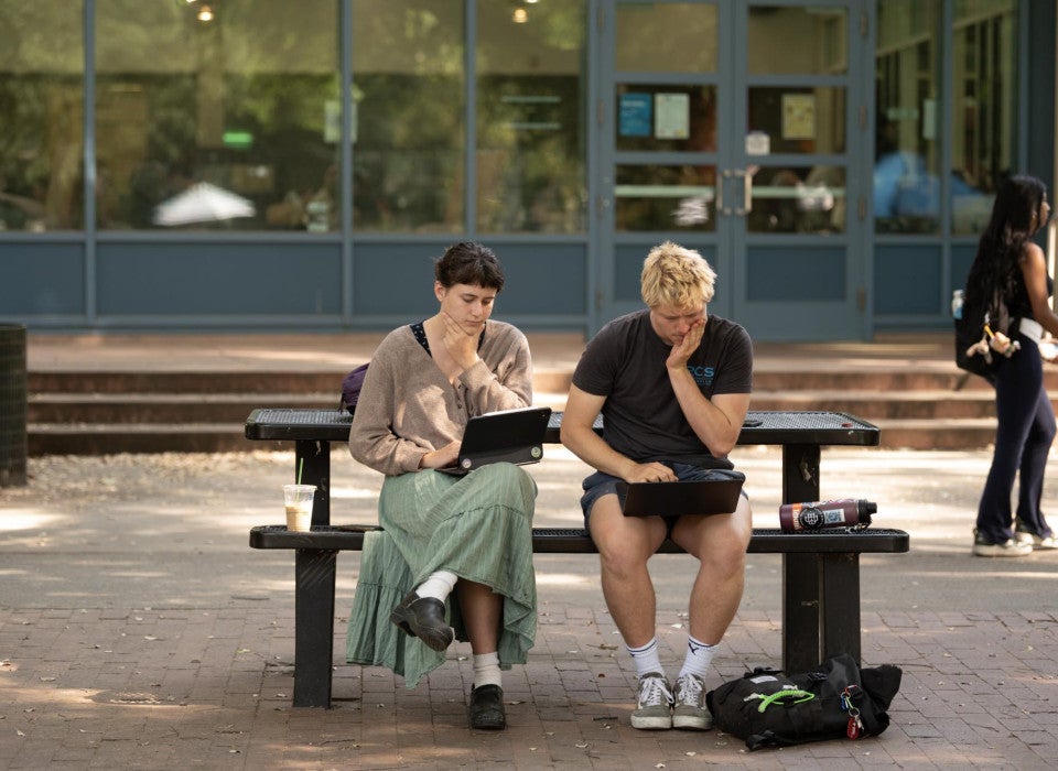 Two students sit at a picnic table outside. They both look down at their laptops in confusion.