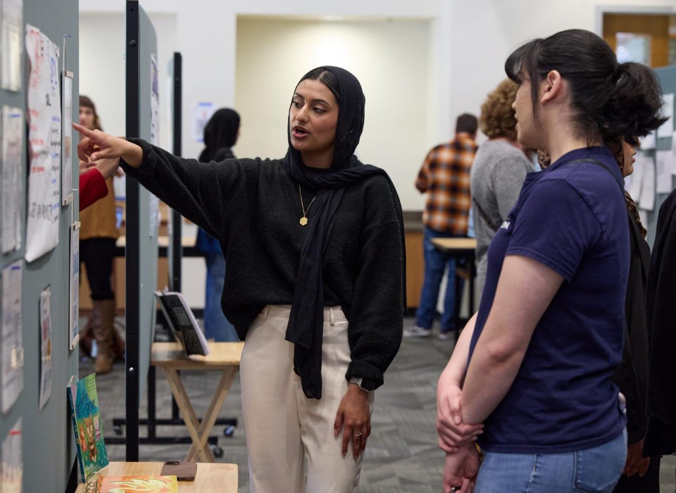 A student in a black shirt, a black hijab, and tan pants points at her research poster while a person in a blue shirt and jeans looks at it.